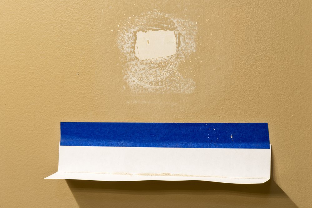 A beige wall showing a small patched area above blue painter’s tape, partially peeled away, highlights recent stucco repair Pasadena work on what was likely a repaired hole or imperfection.
